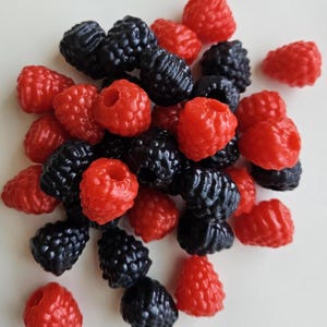 May include: A close-up shot of a pile of red raspberries and black blackberries. The berries are clustered together, with the red raspberries scattered among the darker blackberries. The berries have a glossy appearance and are set against a white background.
