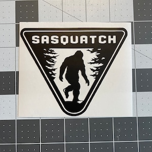 May include: Black and white sticker with a triangular shape. The sticker features a silhouette of a sasquatch walking through a forest. The text "SASQUATCH" is printed in white at the top of the sticker.