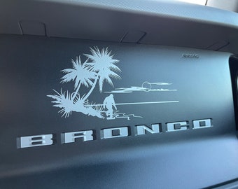 May include: A black dashboard with a white silhouette of palm trees, a sunset, and a figure walking on a beach. The word "BRONCO" is written in white letters below the image. The word "AIRBAG" is printed in black letters on the dashboard.