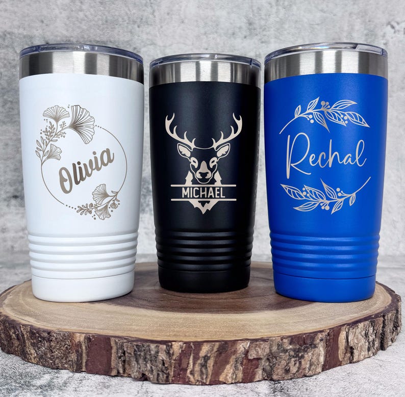 May include: Three stainless steel tumblers with personalized engravings. The white tumbler has a floral design and the name "Olivia". The black tumbler has a deer silhouette and the name "Michael". The blue tumbler has a floral design and the name "Rachel".