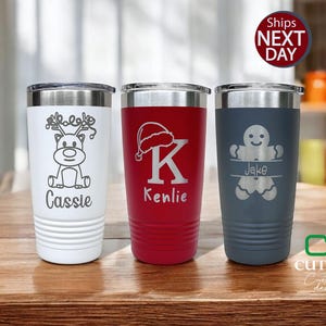 May include: Three personalized stainless steel tumblers with Christmas-themed designs. The tumbler on the left features a reindeer with the name "Cassie" engraved below. The middle tumbler has a Santa hat and the letter "K" with the name "Kenlie" engraved below. The tumbler on the right has a gingerbread man with the name "Jake" engraved below.