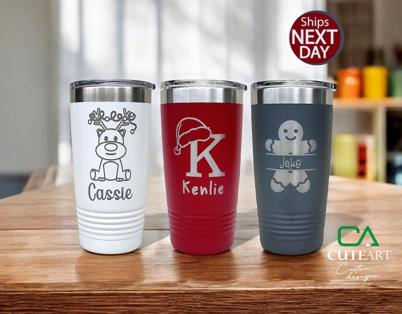 May include: Three personalized stainless steel tumblers with Christmas-themed designs. The tumbler on the left features a reindeer with the name "Cassie" engraved below. The middle tumbler has a Santa hat and the letter "K" with the name "Kenlie" engraved below. The tumbler on the right has a gingerbread man with the name "Jake" engraved below.