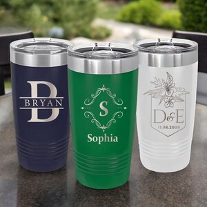 May include: Three stainless steel tumblers with personalized engravings. The tumbler on the left is navy blue with the letter "B" and the name "Bryan" engraved on it. The tumbler in the middle is green with a decorative design and the name "Sophia" engraved on it. The tumbler on the right is white with a floral design and the initials "D&E" and the date "11.08.2009" engraved on it.
