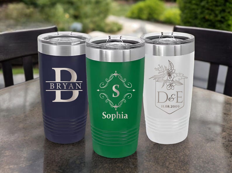 May include: Three stainless steel tumblers with personalized engravings. The tumbler on the left is navy blue with the letter "B" and the name "Bryan" engraved on it. The tumbler in the middle is green with a decorative design and the name "Sophia" engraved on it. The tumbler on the right is white with a floral design and the initials "D&E" and the date "11.08.2009" engraved on it.