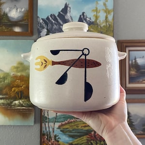 May include: Cream-colored ceramic casserole dish with lid and side handles. The dish features a brown, yellow, and black utensil design. The background includes several framed landscape paintings.