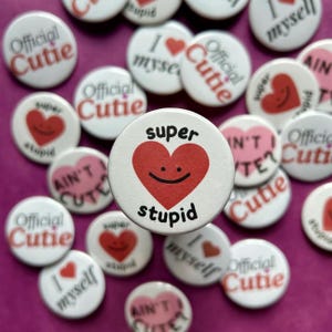 May include: A collection of colorful round pins with various messages, including "Official Cutie", "Super Stupid", "Ain't I Cute?" and "I Love Myself". The pins feature a red heart with a smiling face and the words "Super Stupid" in white.