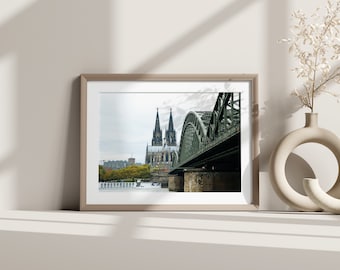May include: A framed photograph of the Cologne Cathedral in Germany, with a bridge in the foreground. The cathedral is a large, gothic structure with two tall spires. The bridge is a steel structure with a green paint job. The photo is taken from a low angle, giving the viewer a sense of the cathedral's height.