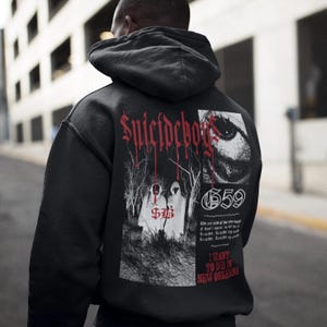 Peut inclure: Sweat à capuche noir avec un motif graphique au dos. Le motif comprend le texte "Suicideboys" en lettres gothiques rouges, un œil stylisé et le texte "I WANT TO DIE IN NEW ORLEANS".