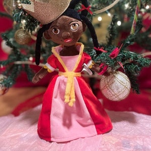 Op de afbeelding: Een pluche pop die een rood-roze jurk draagt met een gele band en een gouden glitterkroon. De pop heeft een bruine huid, zwart haar en bruine ogen. De pop staat voor een kerstboom.