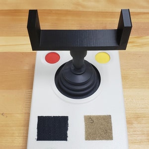 Peut inclure: Un joystick de commande blanc avec une poignée et une base noires. Le contrôleur a deux boutons rouges et jaunes, un joystick noir et deux patchs de velcro noirs. Le contrôleur a un autocollant vert avec le texte "OPTIMAX" et un autocollant bleu avec une coche et le texte "Simply Works".