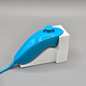 Puede incluir: Un mando Nintendo Wii Remote Plus azul con un soporte de carga blanco. El mando está conectado al soporte con un cable negro.