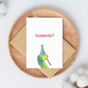 Könnte beinhalten: Eine weiße Grußkarte mit einem grünen Schlange-Motiv, die einen blauen Partyhut mit gelben Punkten trägt. Der Text Sssurprise! ist in Rot über der Schlange gedruckt. Die Karte liegt auf einem braunen Umschlag auf einem runden Holzteller.