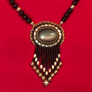 May include: A gold-toned necklace with a large oval pendant featuring a gray stone. The pendant is surrounded by a decorative frame and hangs below a beaded chain with black and gold beads. The necklace has a star-shaped charm at the bottom.