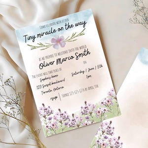 Peut inclure: Une invitation de fête prénatale avec un fond aquarelle représentant des fleurs roses et violettes. Le texte indique "Venez célébrer avec nous notre Petit miracle en route" et "Alors que nous nous préparons à accueillir dans le monde Oliver Marco Smith". L'invitation comprend la date, l'heure et le lieu de l'événement.
