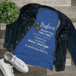 May include: A blue t-shirt with the word "Makers" and inspirational text in white, including "dream big, take risks, show courage." The shirt is paired with a denim jacket and white sneakers, set against a wooden background.