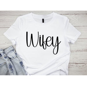 May include: White t-shirt with the word "Wife" written in black cursive script.