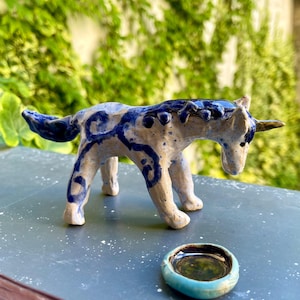 Może przedstawiać: Ceramiczna figurka jednorożca z niebiesko-białą glazurą. Jednorożec ma długi róg i małą, okrągłą, turkusową miseczkę przed sobą. Figurka jest ręcznie robiona i ma unikalny, artystyczny design.