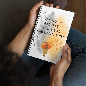 May include: A spiral bound notebook with a white cover and a motivational quote: "It's not a bad day... only bad circumstances". The quote is written in black text. The cover also features a cartoon illustration of a person holding an umbrella in the rain. The background is a watercolor wash of gray and yellow.
