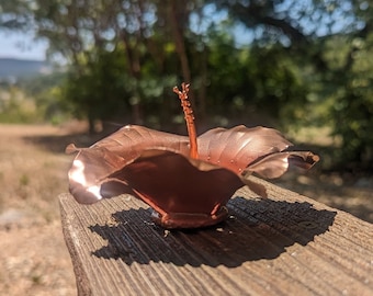 Könnte beinhalten: Eine kupferfarbene Metallblumenskulptur mit einem zarten Stiel und Blütenblättern. Die Skulptur steht auf einer Holzoberfläche.