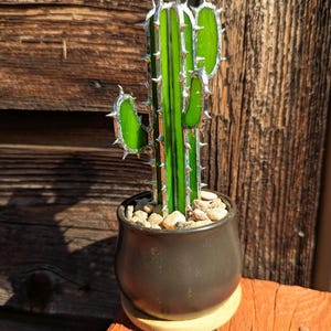 May include: A decorative glass cactus in a black ceramic pot. The cactus features bright green glass segments with silver-colored edges and spiky details. The pot sits on a small wooden coaster, filled with small pebbles.