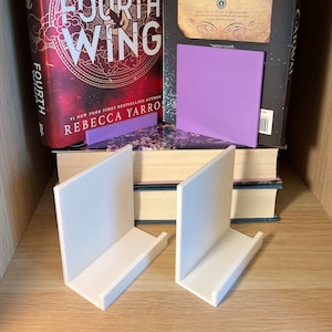 May include: Two white bookends with a curved design, sitting on top of a stack of books. The top book is a fantasy novel titled "Fourth Wing" by Rebecca Yarros. The book is red with gold lettering and a black and white illustration of a bird. The second book is black with gold lettering and a quote about the book. The quote is "A magnificent debut - a spellbinding tale of love, sacrifice, and hope."