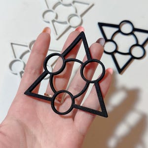 May include: Black geometric shapes with a triangle and circle design. The shapes are held in a hand, with other similar white shapes in the background. The shapes have a modern, minimalist aesthetic.