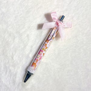 Peut inclure: Un stylo décoratif avec un corps transparent orné d'un motif rose et blanc d'ours de dessin animé. Le stylo a une pointe argentée et est orné d'un nœud en ruban rose. Un manchon en crochet blanc recouvre la base du stylo.