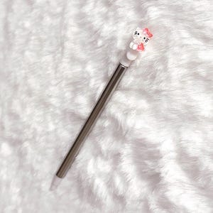 May include: A silver stylus pen with a white tip and a decorative Hello Kitty figure at the top. The figure is white with a pink bow and dress. The pen is shown on a white, fluffy surface.