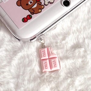 May include: A clear phone case featuring a sticker of a brown bear and strawberries, with a light pink, bitten chocolate bar charm attached by a silver lobster clasp. The charm is embossed with the word MILK on three of its rectangular segments.
