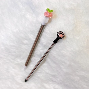 May include: Two metal stylus pens with decorative toppers. One stylus features a pink peach fruit with a green leaf topper, while the other stylus features a black cat paw print design with a pink center pad. Both pens have silver metallic barrels.