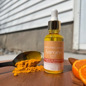 May include: A glass bottle of Aurora Turmeric & Vitamin C Facial Serum, a skincare product, with a gold lid and dropper. The bottle is sitting on a wooden surface with a spoon of turmeric powder and orange slices.