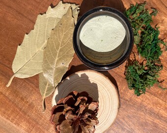 May include: A small glass jar with a lid filled with a light green powder. The jar is sitting on a wooden surface with a pine cone, dried leaves, and green moss.
