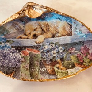 May include: A golden seashell with a painted image of a sleeping puppy, flowers, and gardening tools. The seashell is a decorative trinket.