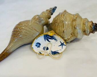 May include: A white ceramic seashell dish with a blue and gold design featuring sea life and a starfish. A pearl necklace is resting inside the dish. Two beige ceramic seashells are in the background.