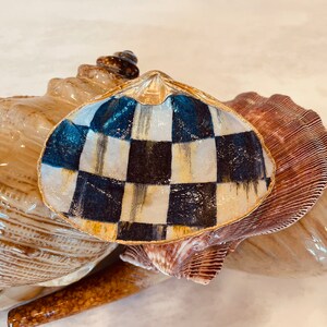 May include: A seashell with a black and white checkered pattern painted on it. The shell is gold-trimmed and sits on top of another seashell.