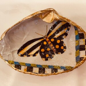 May include: A gold-rimmed seashell dish with a painted butterfly design. The butterfly has black and white stripes, yellow and black polka dots, and a brown body.