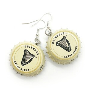 May include: A pair of earrings made from Guinness beer bottle caps. The caps are cream colored with black lettering that reads "GUINNESS ESTD 1759 EXTRA STOUT" and a black harp logo. The earrings are attached to silver hooks.