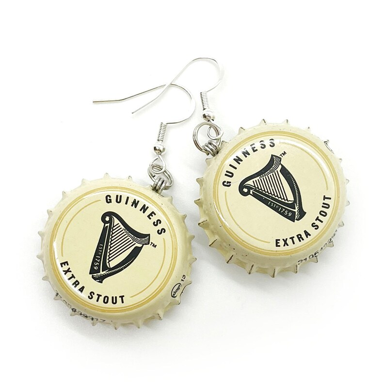 May include: A pair of earrings made from Guinness beer bottle caps. The caps are cream colored with black lettering that reads "GUINNESS ESTD 1759 EXTRA STOUT" and a black harp logo. The earrings are attached to silver hooks.