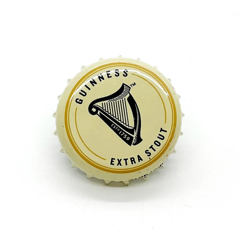May include: A Guinness Extra Stout bottle cap. The cap has a cream-colored center with the Guinness logo, a black harp, and the text "GUINNESS" and "EXTRA STOUT". The rim is metallic gold.