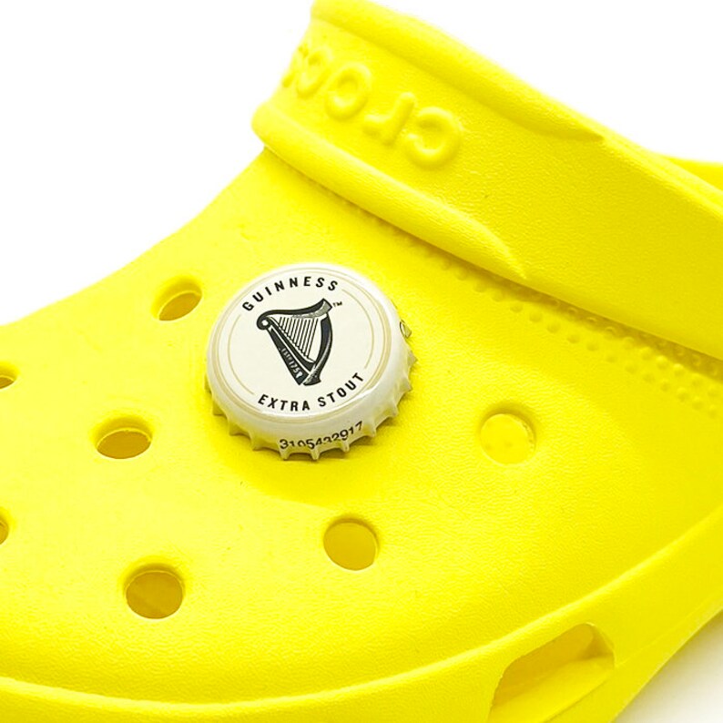 May include: A yellow clog with a Guinness Extra Stout bottle cap charm. The bottle cap is white with black lettering and a harp logo.