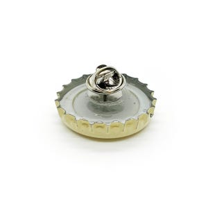 May include: A bottle cap pin with a silver pin back. The pin features a silver-colored pin back attached to a bottle cap with a silver rim and a light-colored interior. The bottle cap has a silver rim and a light-colored interior.