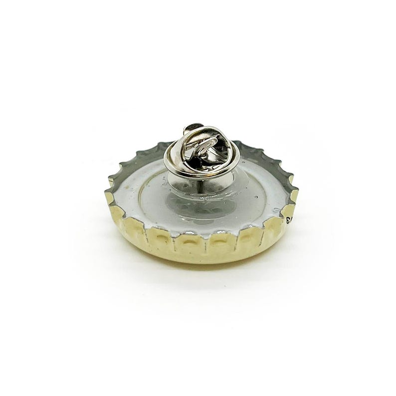 May include: A bottle cap pin with a silver pin back. The pin features a silver-colored pin back attached to a bottle cap with a silver rim and a light-colored interior. The bottle cap has a silver rim and a light-colored interior.