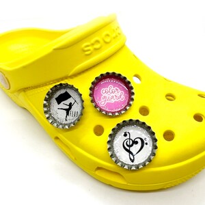 May include: Three colourful bottle cap charms with glitter. The charms feature a black and white silhouette of a colour guard with a flag, a pink background with the text "colour guard", and a black and white musical note with a heart.