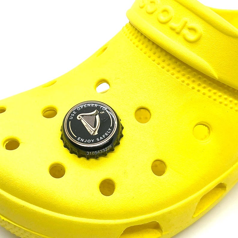 May include: A bright yellow Crocs shoe with a bottle cap charm. The charm is black and silver, featuring the text "USE OPENER TO ENJOY SAFELY" and a harp logo. The charm is attached to the shoe.