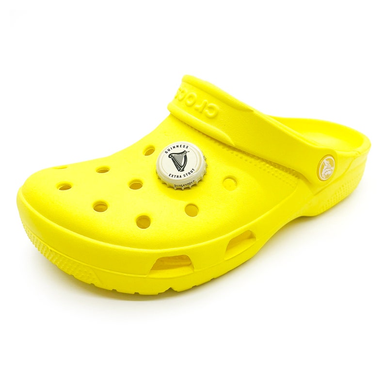 May include: A yellow pair of clogs with a Guinness Extra Stout bottle cap charm. The charm features the Guinness harp logo and the words "Guinness Extra Stout".
