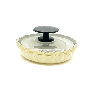 May include: A white bottle cap with a black plastic button on top. The bottle cap has a gold rim and a textured surface.