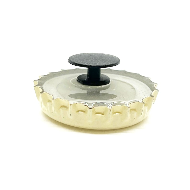 May include: A white bottle cap with a black plastic button on top. The bottle cap has a gold rim and a textured surface.
