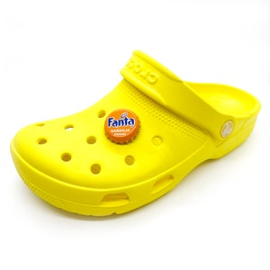 May include: Bright yellow Crocs shoe with a Fanta bottle cap charm. The charm is orange and features the Fanta logo and the words "NARANJA ORANGE". The shoe has multiple holes and a strap.
