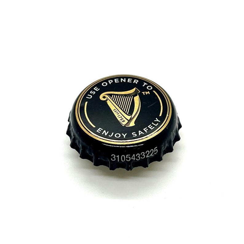 May include: A black bottle cap with gold lettering and a gold harp design. The text on the cap reads "USE OPENER TO ENJOY SAFELY" and "EST 1759". The cap also has the number "3105433225" printed on it.