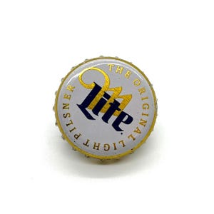 May include: A Miller Lite beer bottle cap. The cap is white with gold trim and features the Miller Lite logo in blue and gold. The text "THE ORIGINAL LIGHT PILSNER" is printed around the edge.